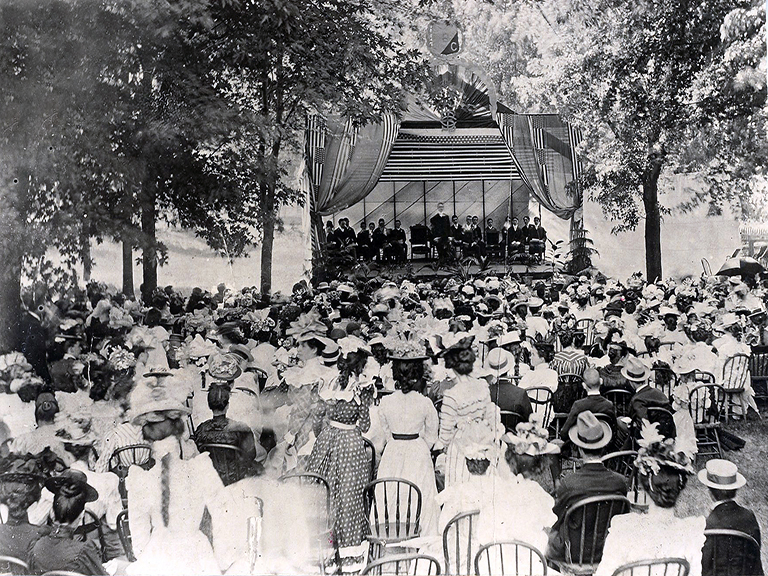 On a small stage with American flag drapery about 2 dozen young men dressed in suits are seated facing a large well-dressed crowd seated in chairs under trees.